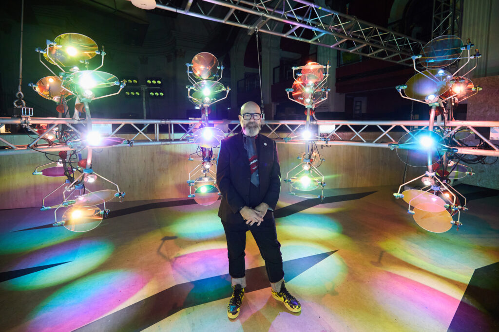 A person in glasses and a suit stands smiling in front of a row of colorful, futuristic light sculptures, casting vibrant reflections on the floor in an indoor venue.