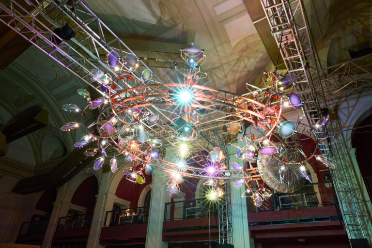 A large, circular, modern chandelier with colorful lights and reflective surfaces is suspended from a metal frame inside a grand hall with high ceilings and arched windows.