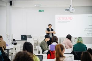 A man stands at the front of a classroom giving a presentation on "Outbound" to students seated at desks, with a projector screen displaying his slides in the background.