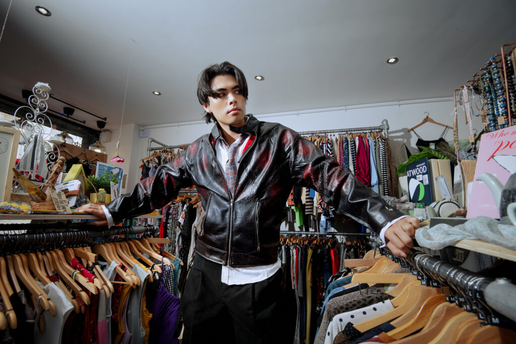 A person wearing a black leather jacket stands confidently in a vintage clothing store, surrounded by racks of colorful clothes and accessories.