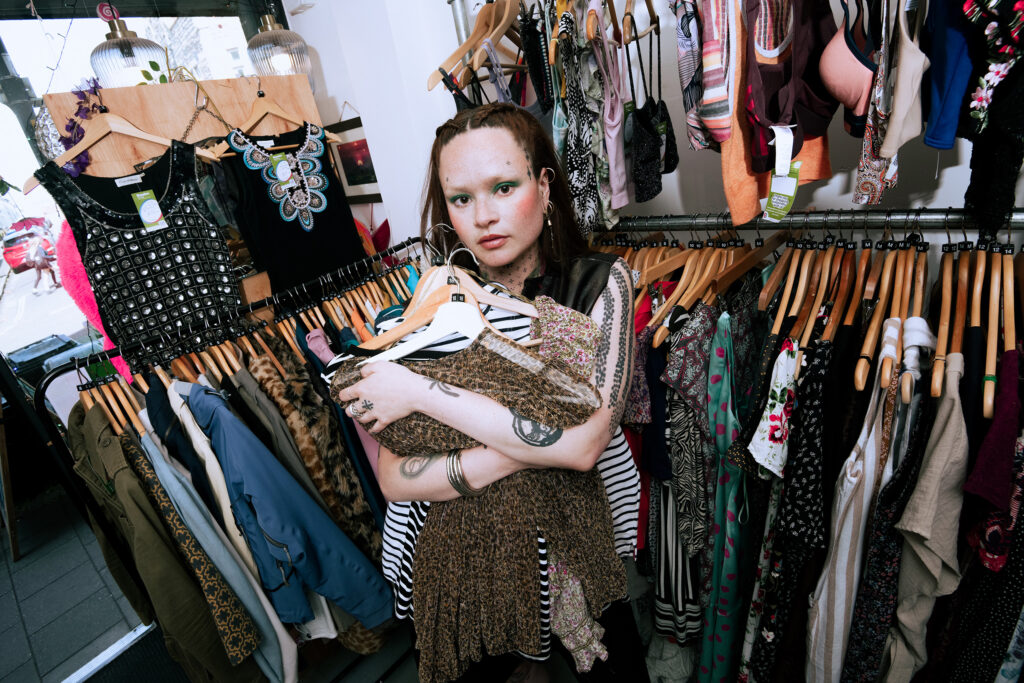 A person with light skin and freckles holds several garments in their arms while standing in a clothing store surrounded by racks of colorful clothes. They look directly at the camera with a neutral expression.