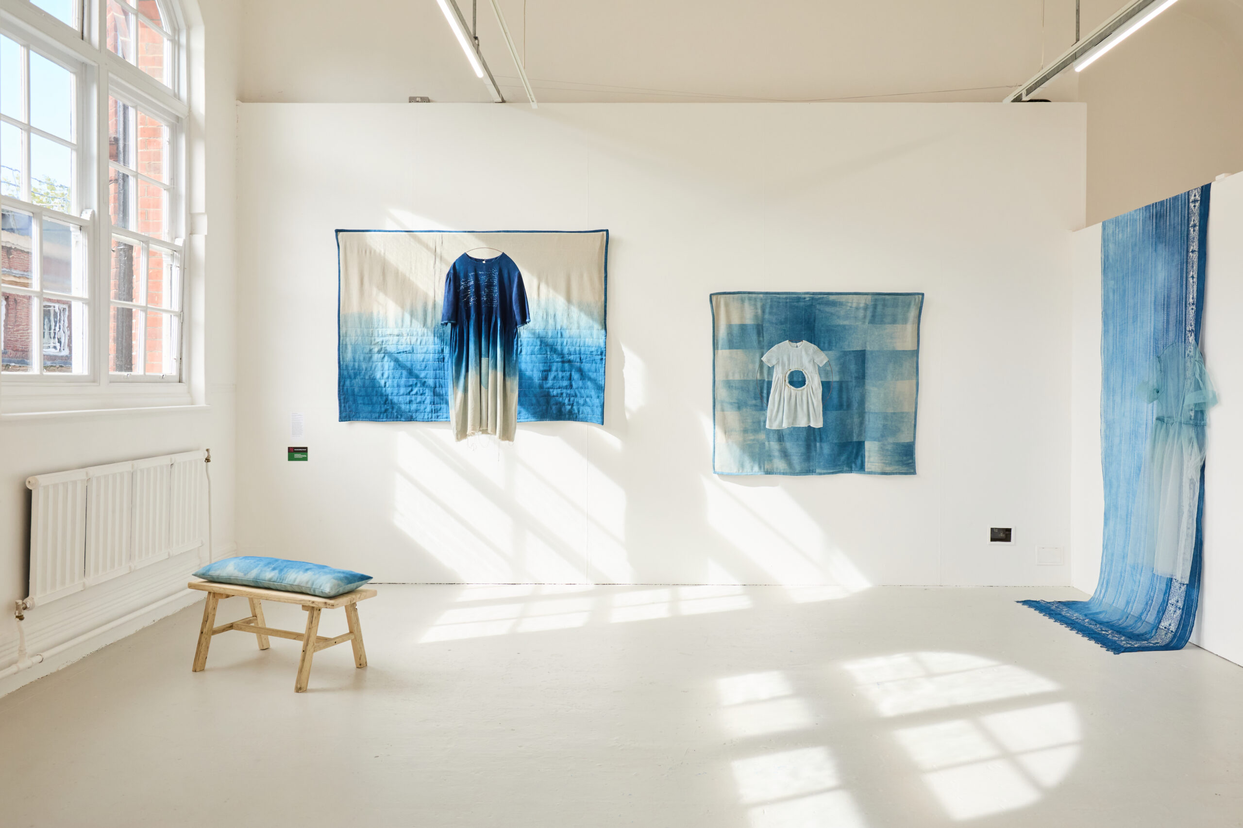 A bright art gallery with white walls displays blue ombre textile art, including a hanging shirt, a small dress, and fabric draped to the floor. A small wooden bench with a blue cushion sits near large windows.