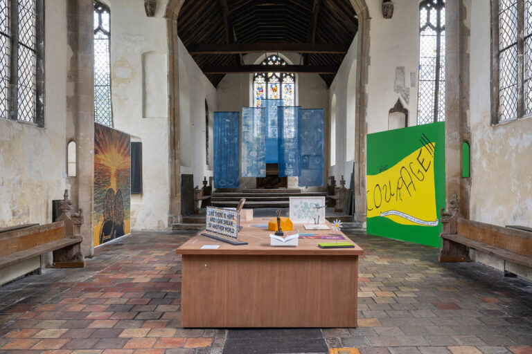 An art installation inside a church features colorful panels, including one with “COURAGE” in bold letters, drawings on a central table, and hanging blue fabric with stained glass windows in the background.