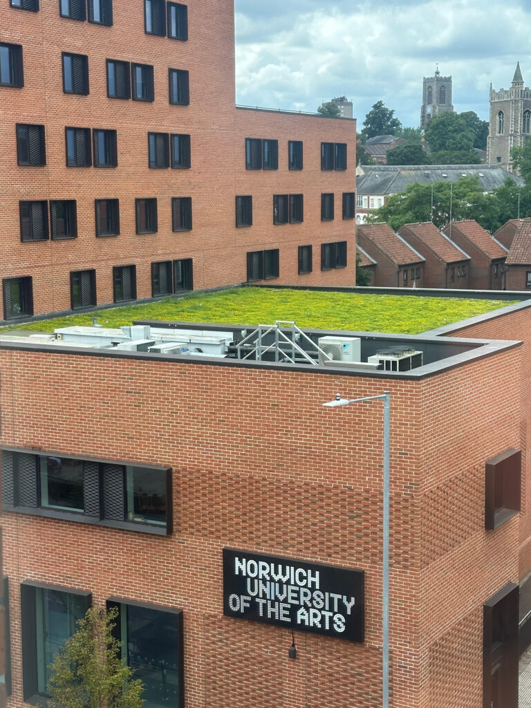 View of a modern red-brick building labeled “Norwich University of the Arts,” with a green rooftop and another building and church visible in the background under a cloudy sky.