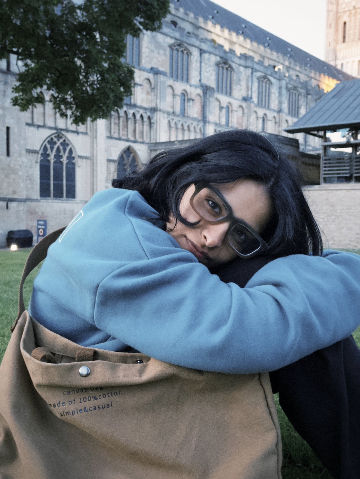 A person with glasses and dark hair sits on grass, hugging their knees and resting their head on their arms. They wear a blue sweatshirt and have a tan canvas bag. Norwich Cathedral is in the background.