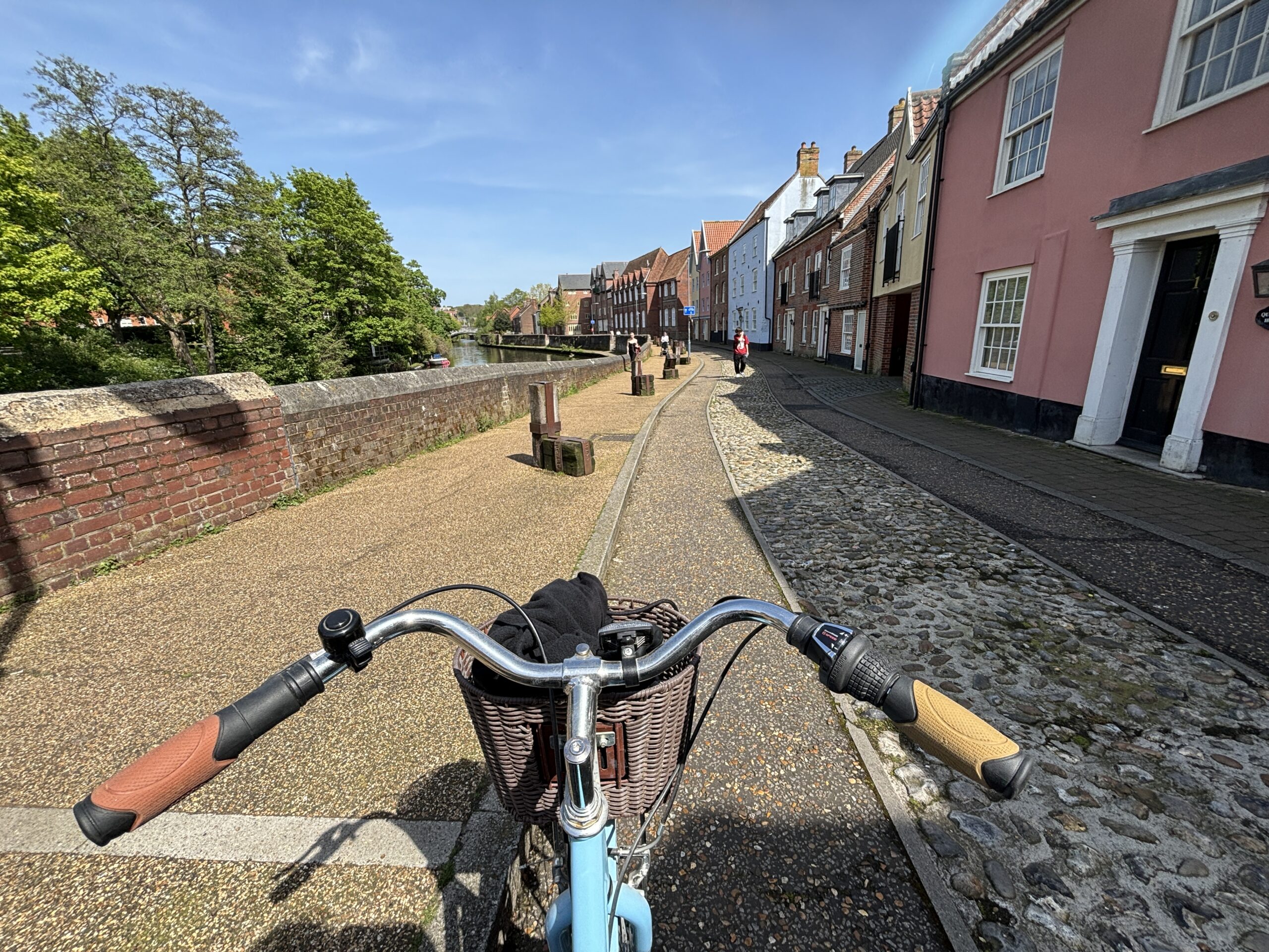 View from a bicycle handlebar on a sunny path beside a river and colourful houses; cobblestone and brick sidewalks with trees and clear blue sky in the background.