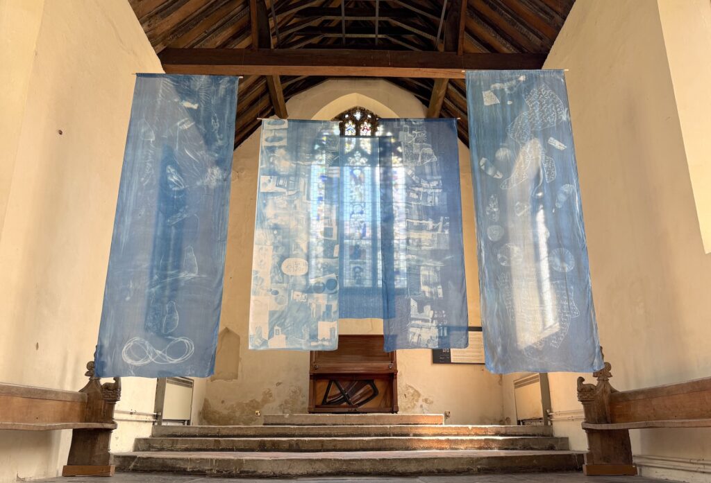 Three large translucent blue fabric banners with white abstract designs hang in front of a stained glass window in a church with wooden pews and a high, wooden-beamed ceiling. Sunlight shines through the window and banners.