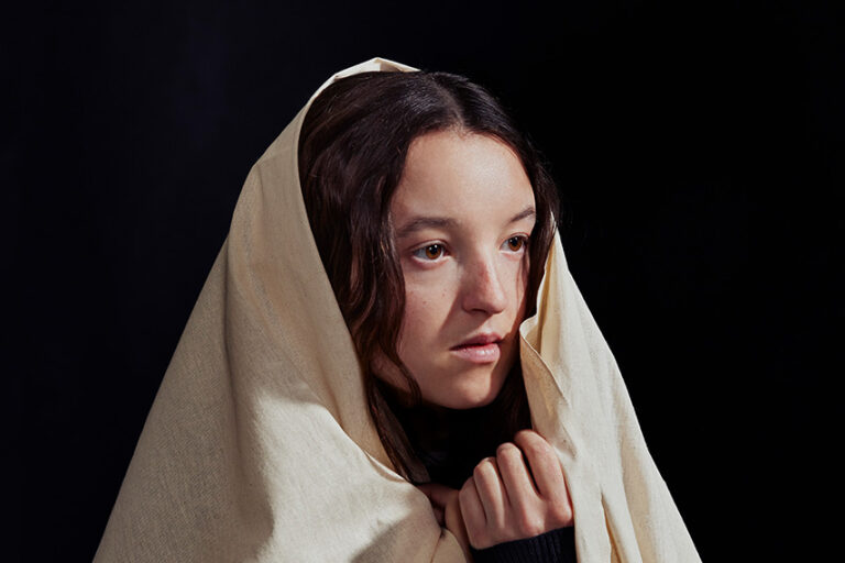 A young person with long brown hair gazes thoughtfully to the side, wrapped in a light beige cloth against a dark background.