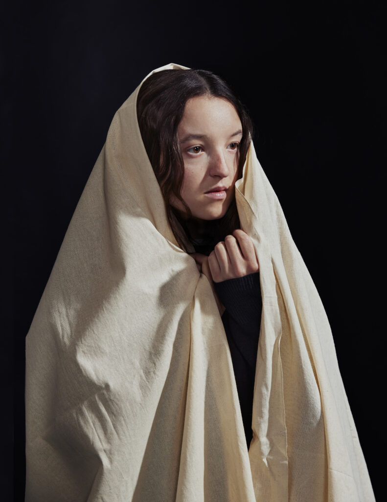 A woman with long brown hair is wrapped in a light beige cloth, covering most of her body and head. She gazes thoughtfully to the side against a dark, plain background.