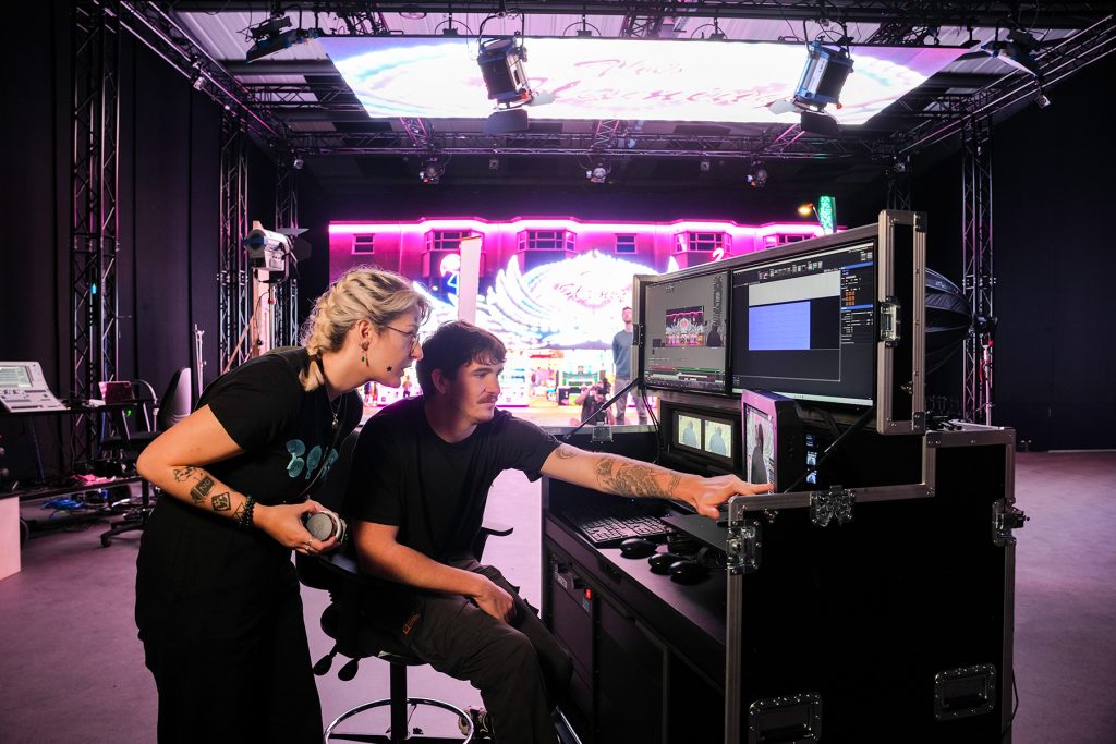 Two people work at a control console with multiple monitors in a stage production setting, reviewing visuals and data, with a brightly lit stage and large screen in the background.