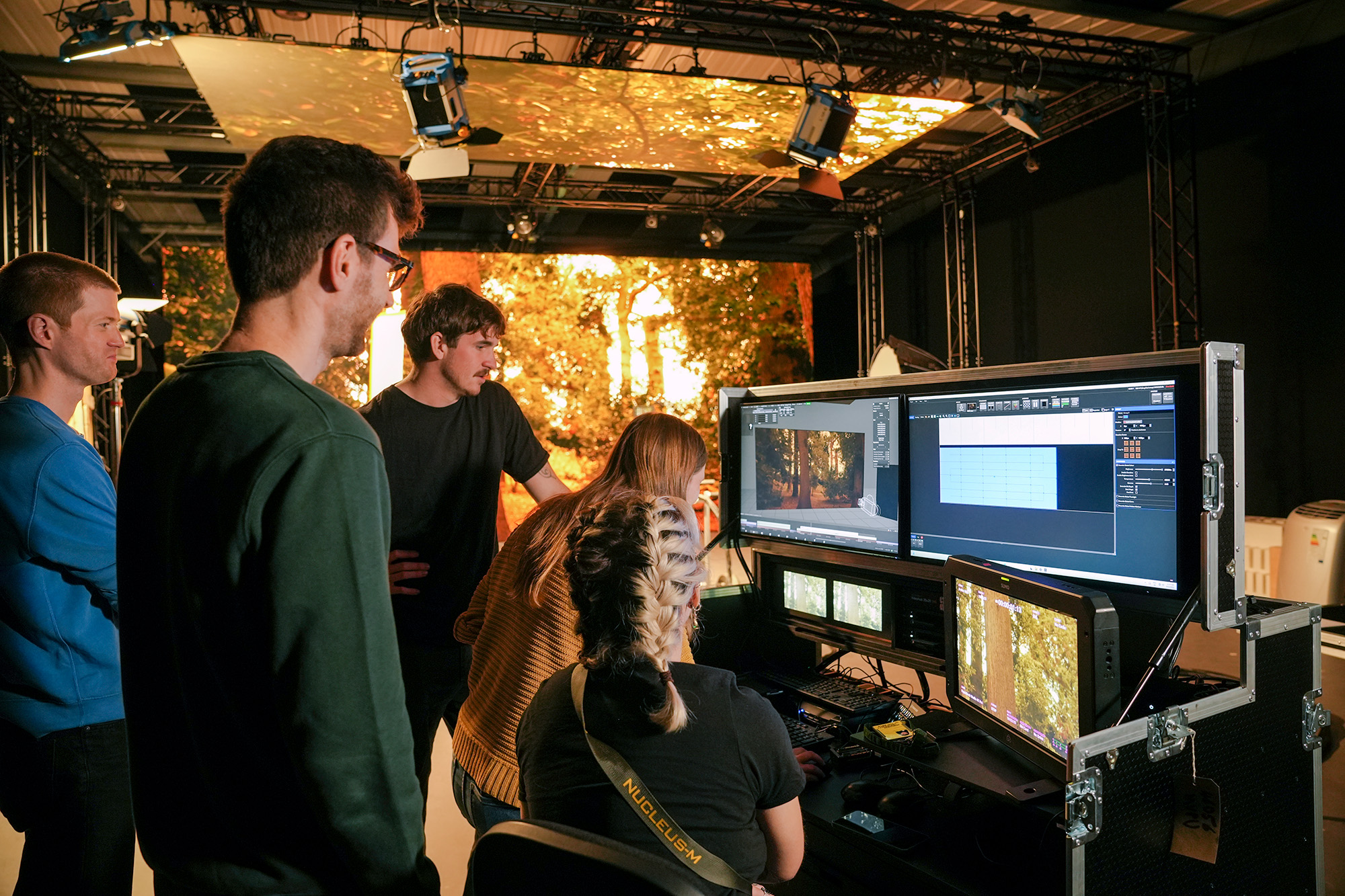 A group of people work together in a studio, gathered around computer monitors displaying digital graphics and a forest scene, with cables and equipment visible in the background.