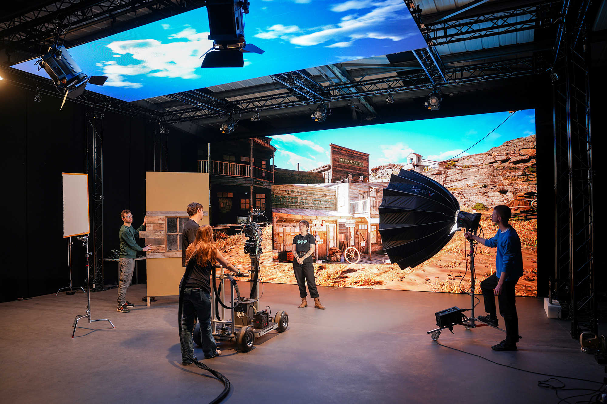 Four people work on a film set with studio lights and cameras in front of a large screen displaying a Western desert town scene, creating the illusion of being outdoors.