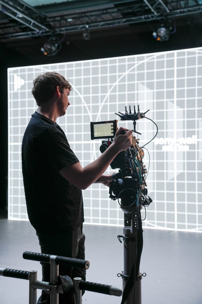 A person operates a professional video camera mounted on a rig in a studio, with a large grid-patterned screen and soft overhead lighting in the background.