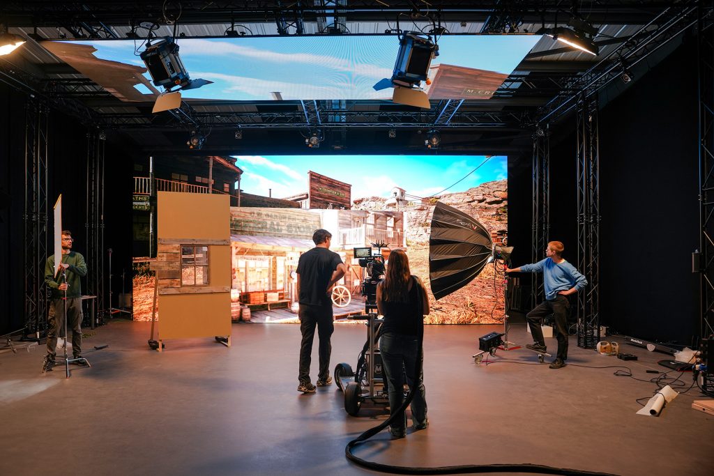 Four people work on a film set with professional cameras, lights, and a large digital screen displaying a Western town backdrop. Equipment and props are arranged around the studio.
