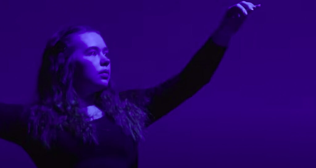 A person with long hair stands with arms raised, illuminated by purple lighting against a dark background—capturing the dramatic, artistic atmosphere of our upcoming art open day.
