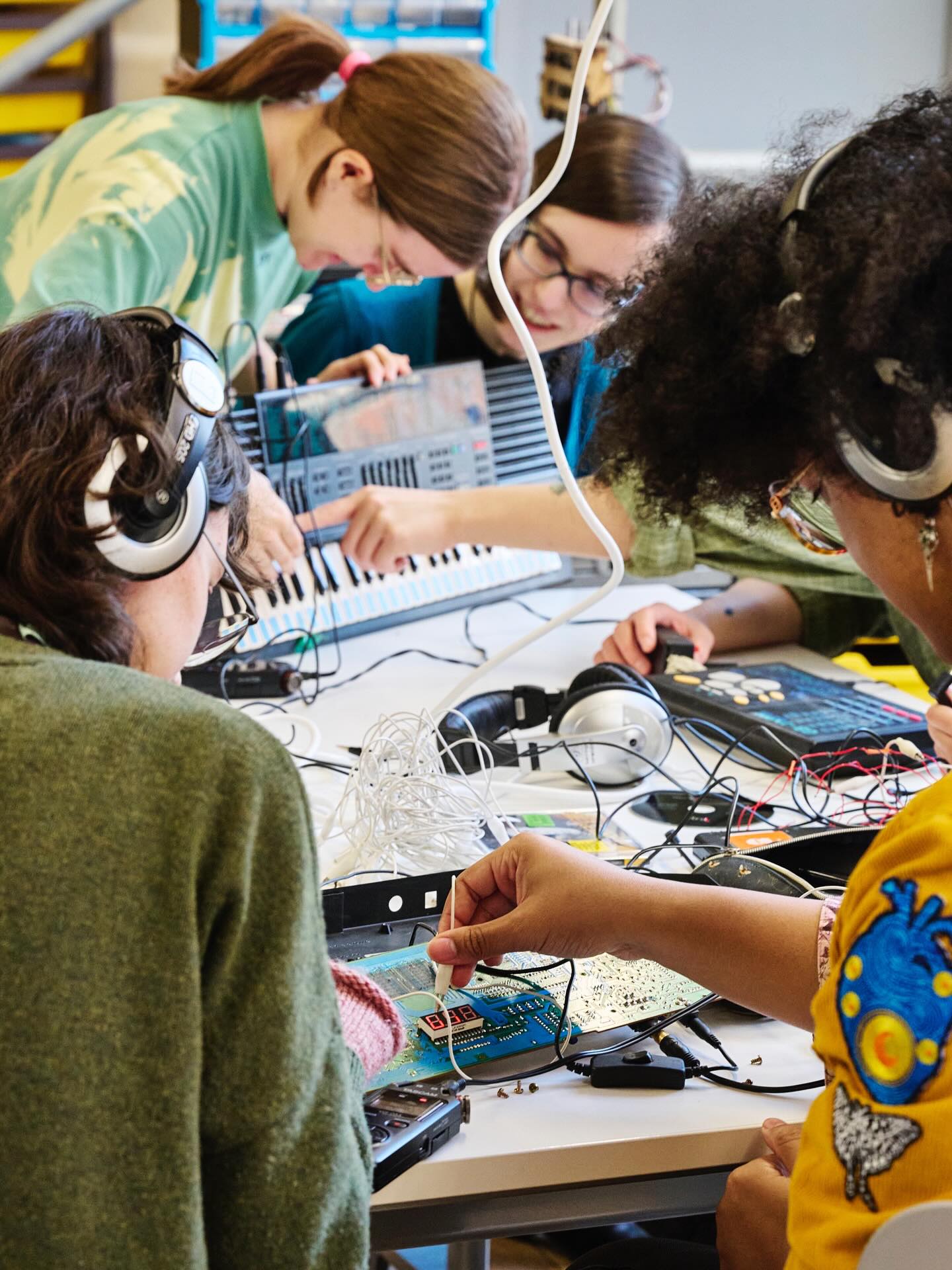 Four people wearing headphones work together on electronic projects at a table, using circuit boards, wires, and a keyboard. They appear focused and engaged in collaborative hands-on activities.