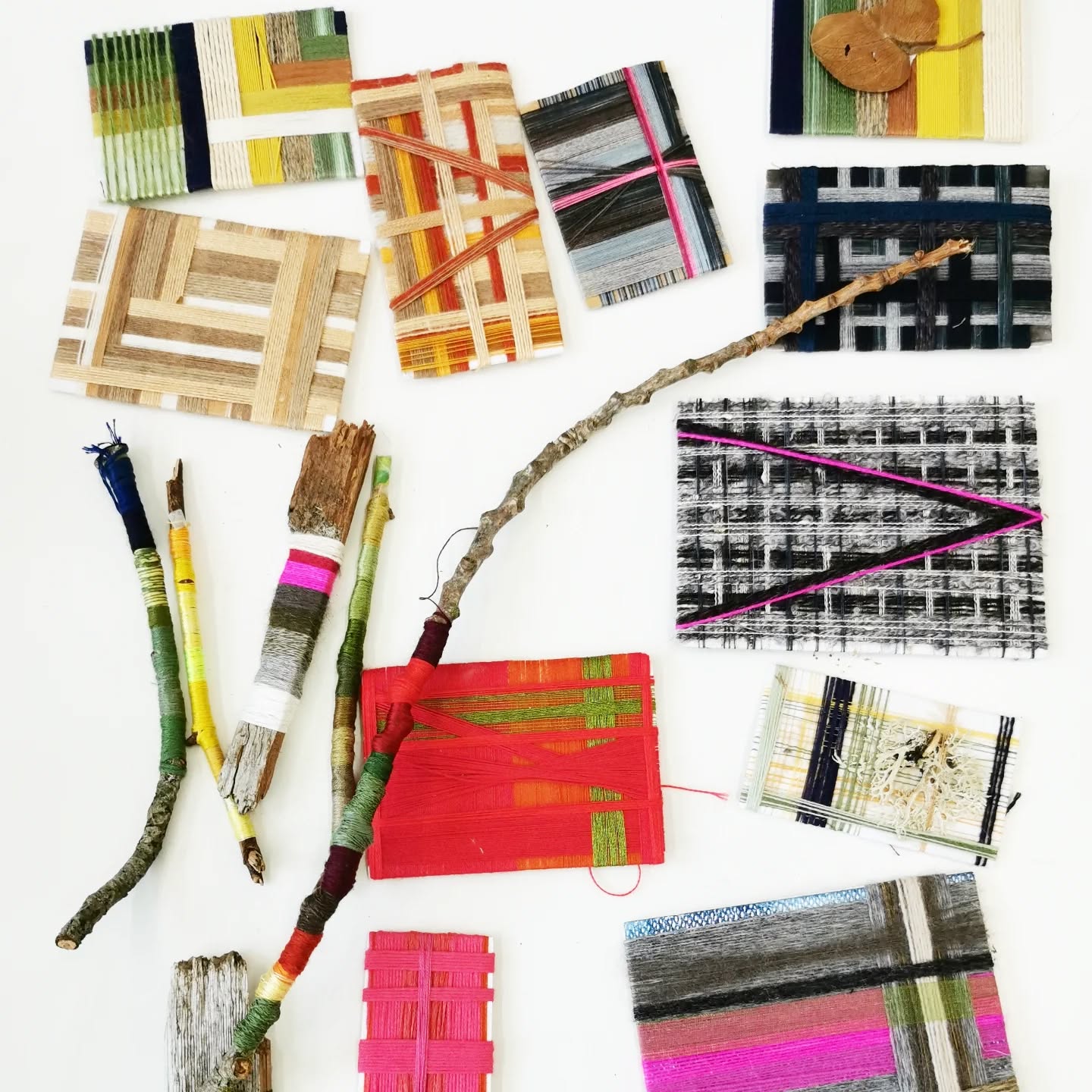 Various pieces of woven art, featuring colorful threads wrapped around cardboard and sticks, are arranged on a white surface. The designs include stripes, checks, and abstract patterns in vibrant colors.