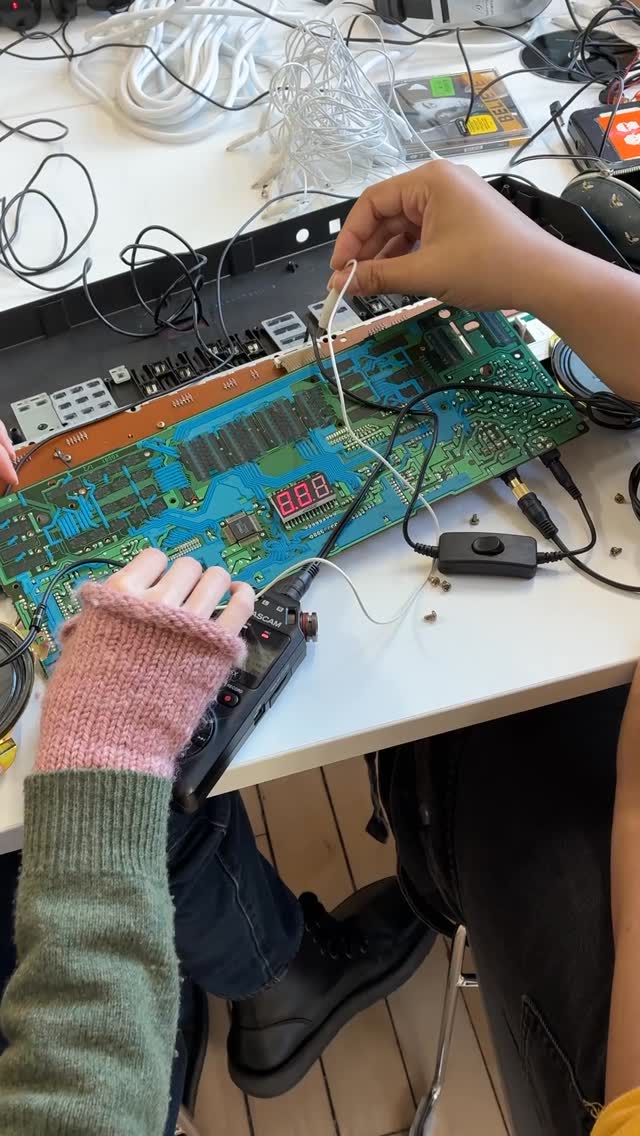 Two people work on an exposed circuit board with wires and electronic components; one wears a pink-and-green fingerless glove, and tools and cables are scattered on the white table.