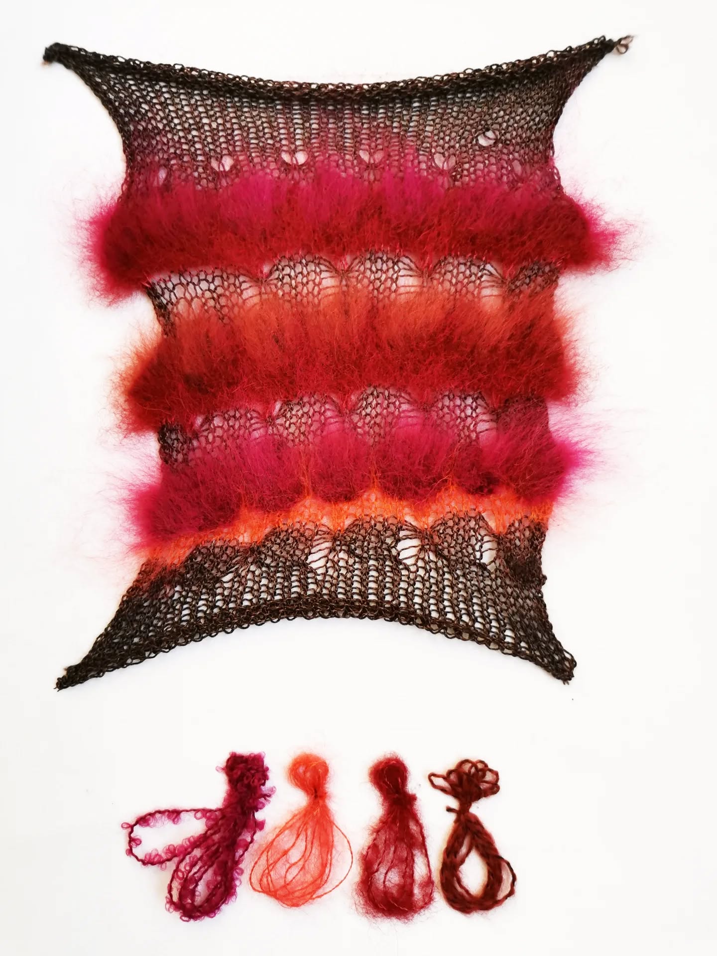 A rectangular swatch of knitted lace in black, red, orange, and pink fuzzy yarns is displayed above four small bundles of matching yarn colors, arranged in a row on a white background.