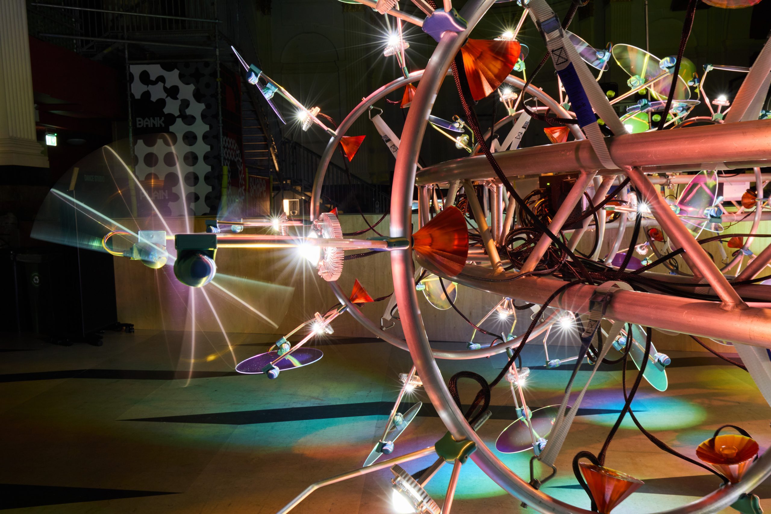 A large, futuristic light sculpture made of metal rods, funnels, and various colorful lights glows brightly in a dark indoor space, creating reflections and starburst effects on the floor.