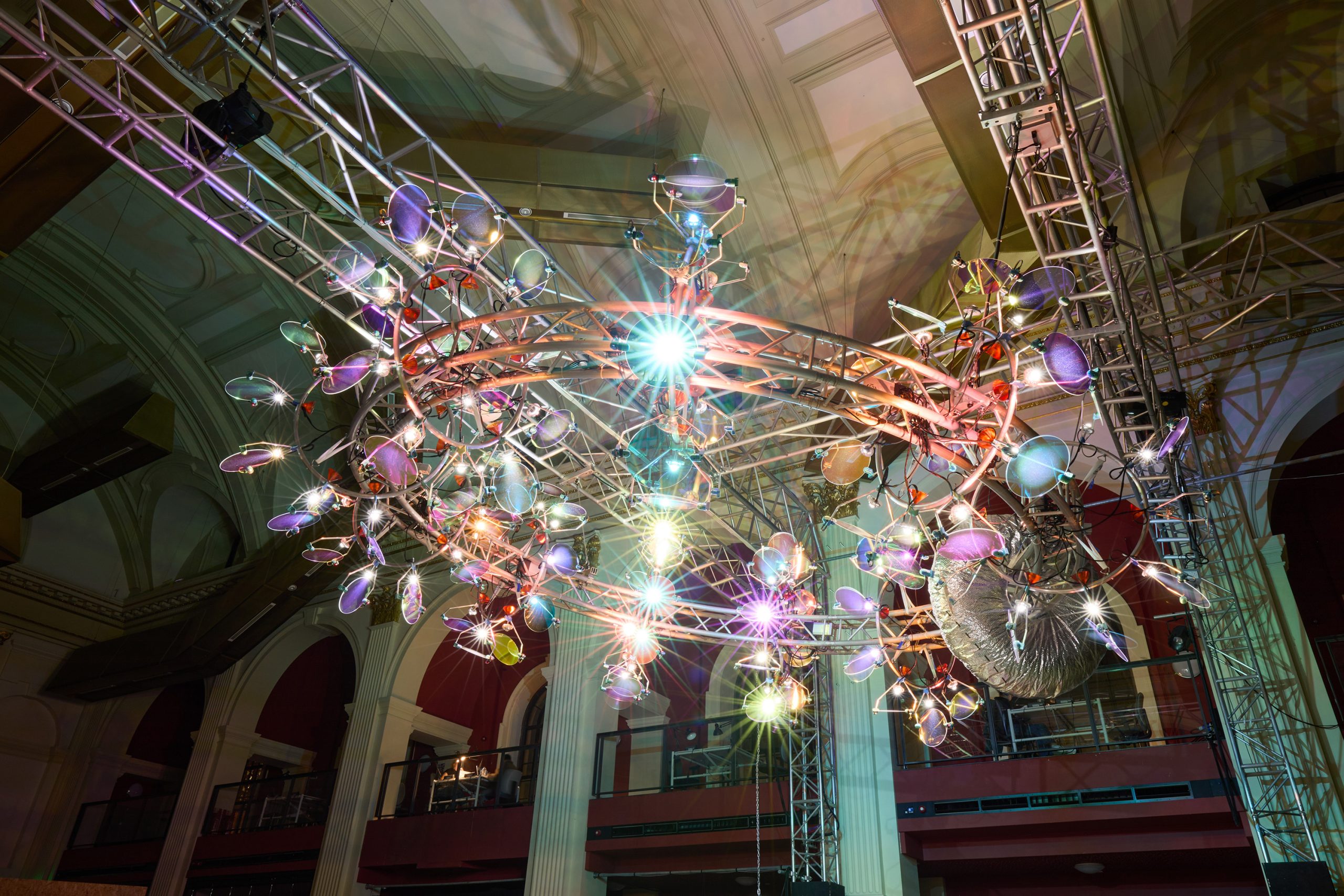 A large, circular, modern chandelier with colorful lights and reflective surfaces is suspended from a metal frame inside a grand hall with high ceilings and arched windows.
