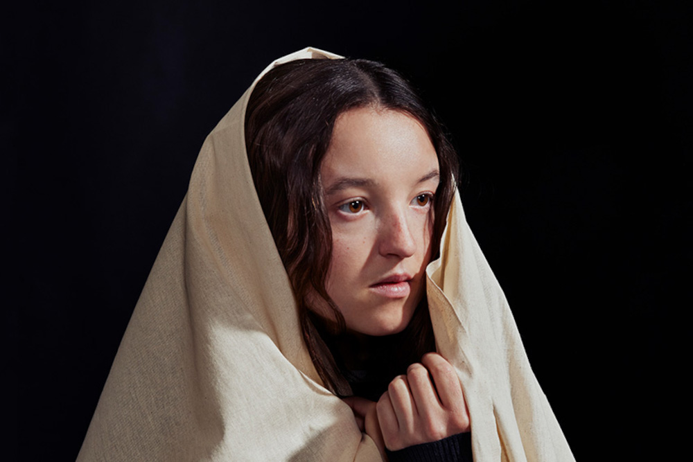 A woman with long brown hair is wrapped in a light beige cloth, covering most of her body and head. She gazes thoughtfully to the side against a dark, plain background.