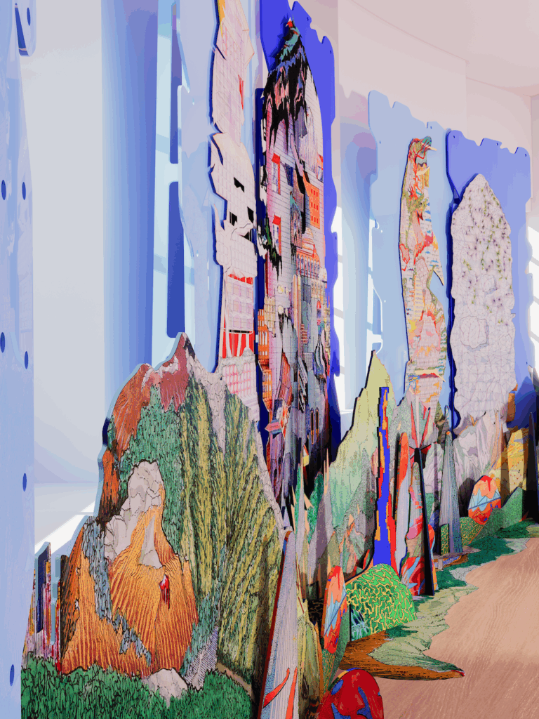Colorful, layered cardboard cutouts resembling abstract mountains and buildings are arranged indoors on a wooden floor, with light streaming in through large windows in the background.