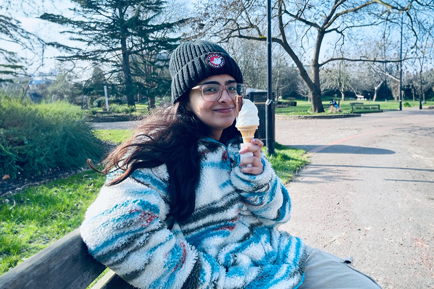 A person wearing glasses, a beanie, and a fuzzy striped jacket sits on a park bench, smiling and holding an ice cream cone. The park is sunny with bare trees and green grass in the background.