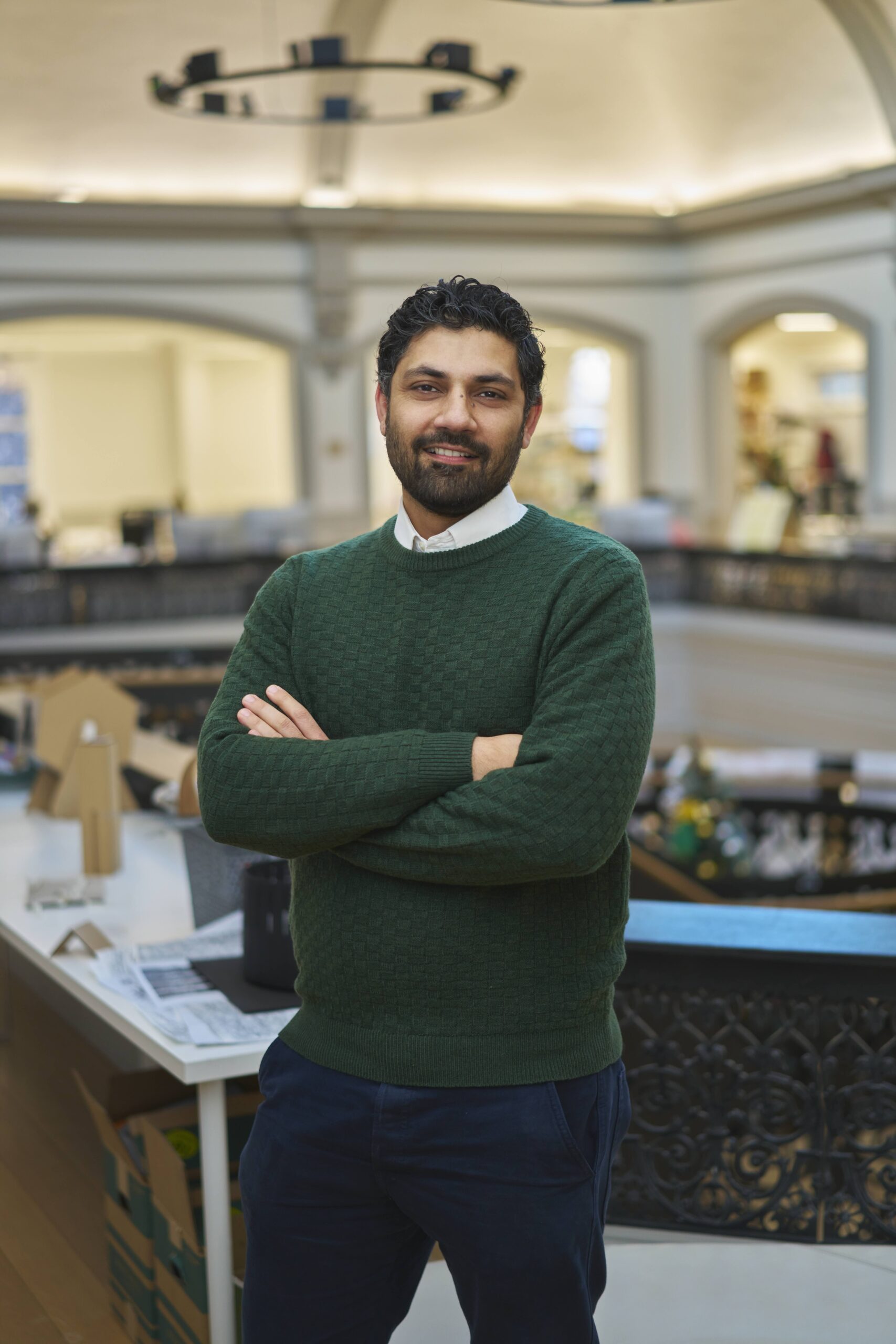 A man with dark hair and a beard, wearing a green sweater and white shirt, stands with arms crossed and smiles in a bright, modern indoor space with tables and decorative railings.