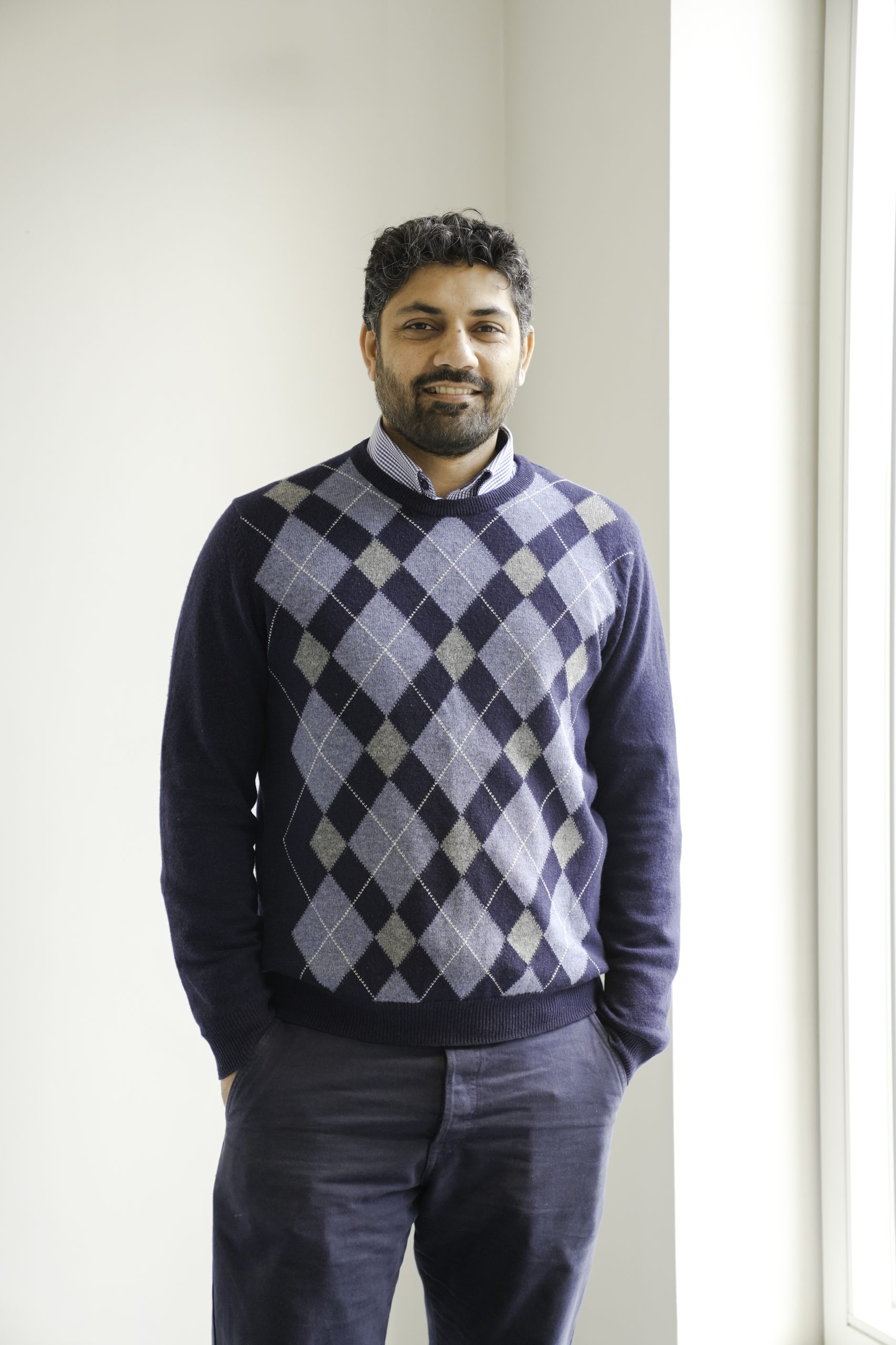A man with dark curly hair and a beard stands by a bright window, wearing a blue argyle sweater over a collared shirt and dark trousers. He is smiling and has his hands in his pockets.