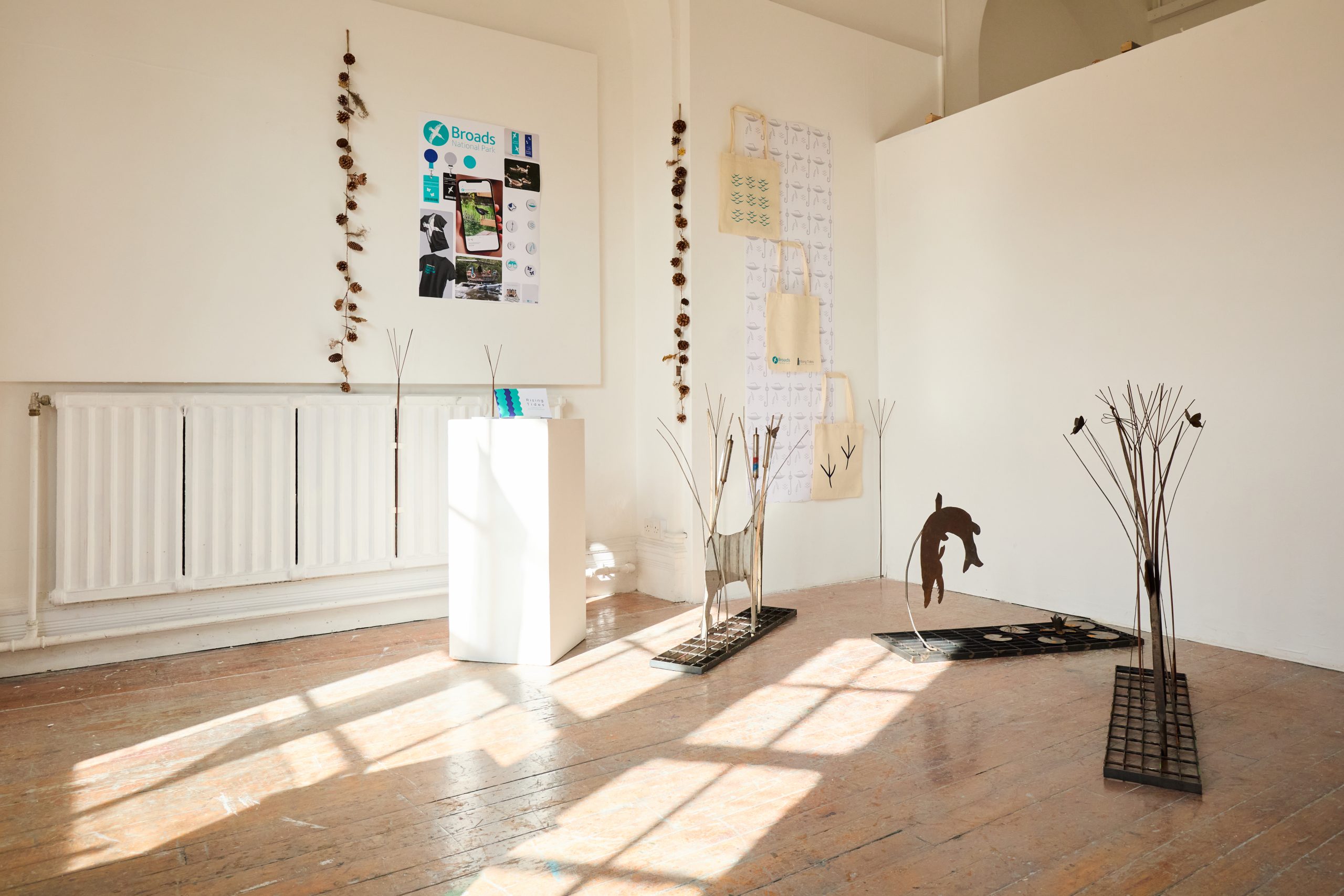 A bright art gallery space with wooden floors, featuring metal animal sculptures, wall hangings and posters, with sunlight streaming through large windows onto the floor.
