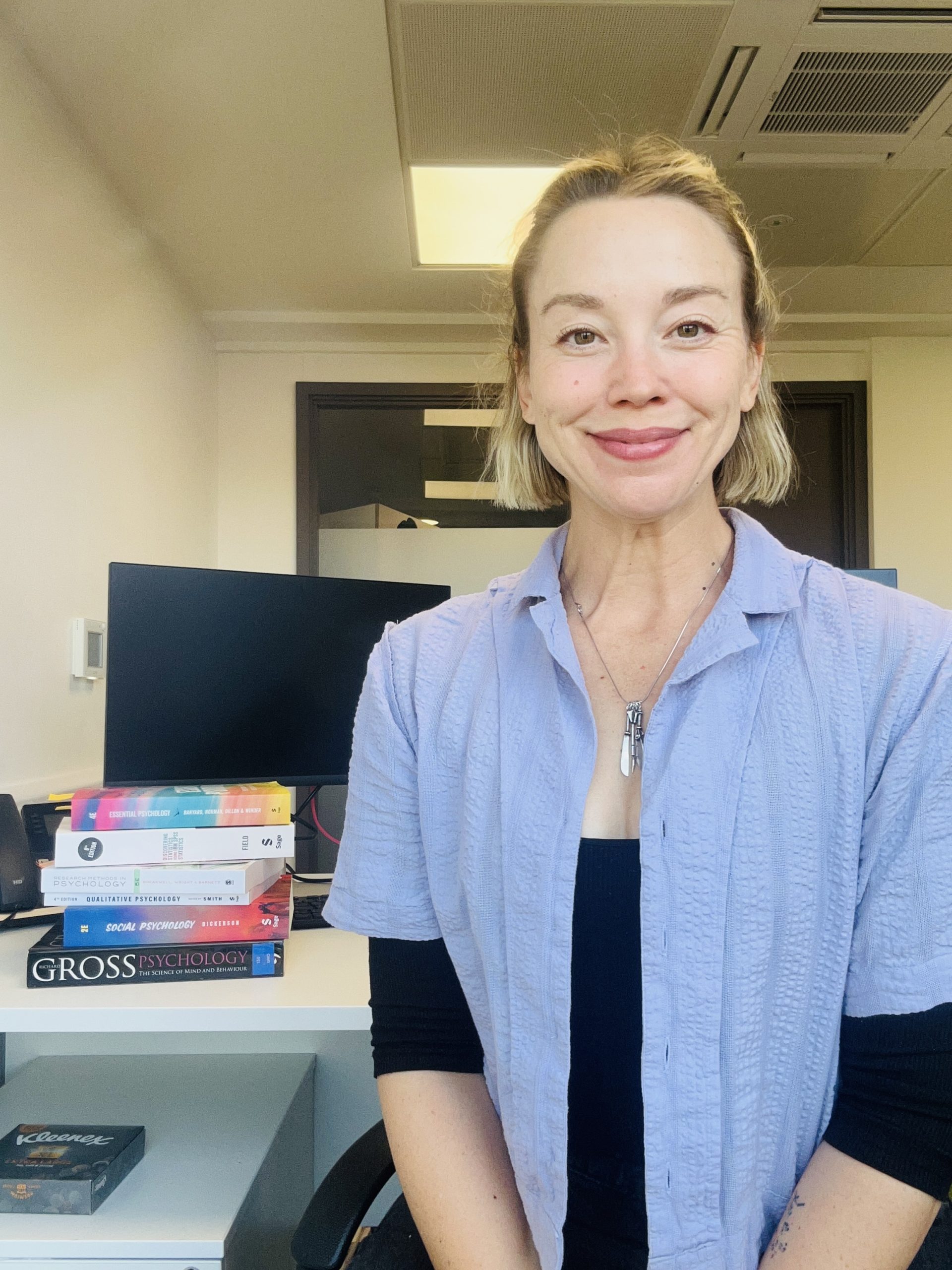 A smiling woman with blonde hair sits in an office with a computer, filing cabinet, and a stack of books on psychology and psychiatry beside her. She wears a light purple shirt over a black top.