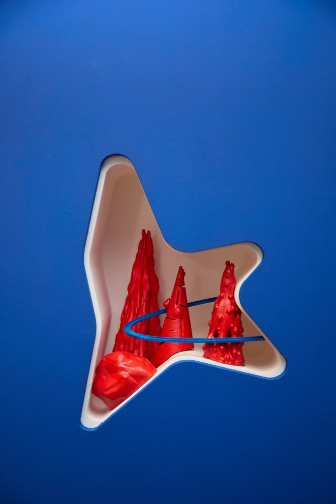 A star-shaped cutout in a blue wall frames red abstract sculptures inside, with a blue curved line crossing in front of them.