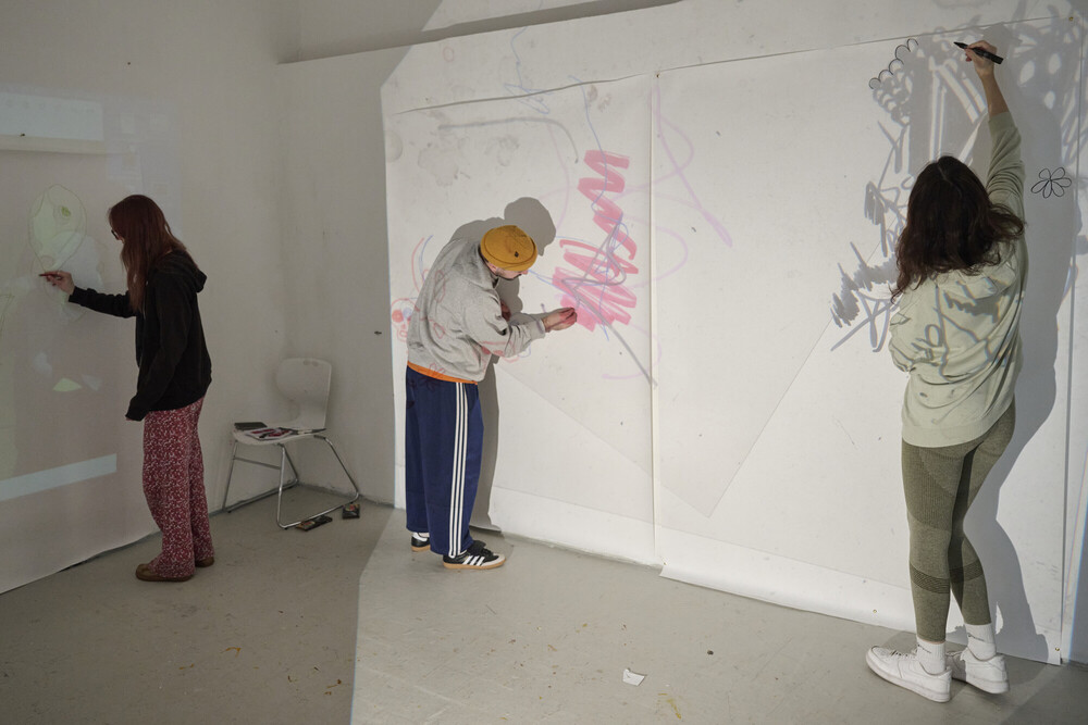 Three people are drawing on a large white wall in an art studio. Two women and one man are using markers to create colorful sketches and shapes. The room is bright and minimal, with art supplies on the floor.