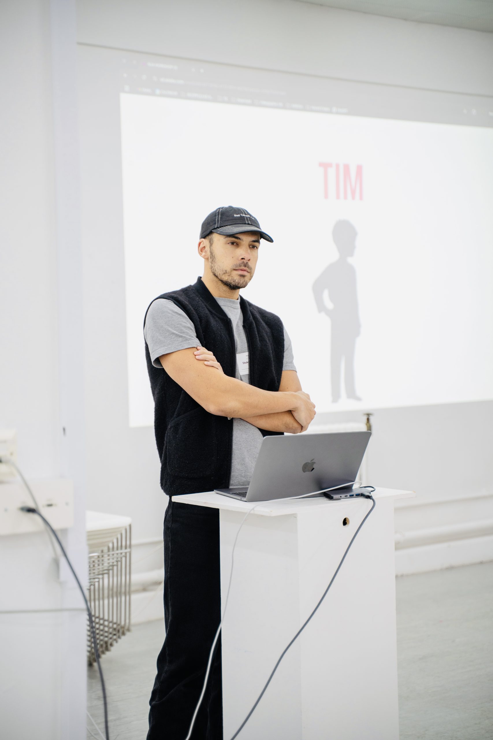 A man in casual attire, wearing a cap and vest, stands with arms crossed behind a podium with a laptop. A presentation slide with the word “TIM” and a silhouette figure is projected behind him.