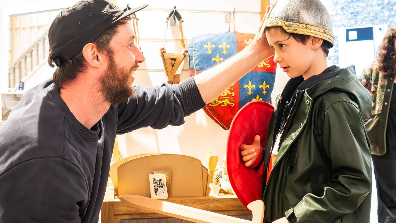 A man kneels and smiles at a child wearing a toy knight helmet and holding a red shield, surrounded by medieval-themed play items. They appear to be playing or at an event.