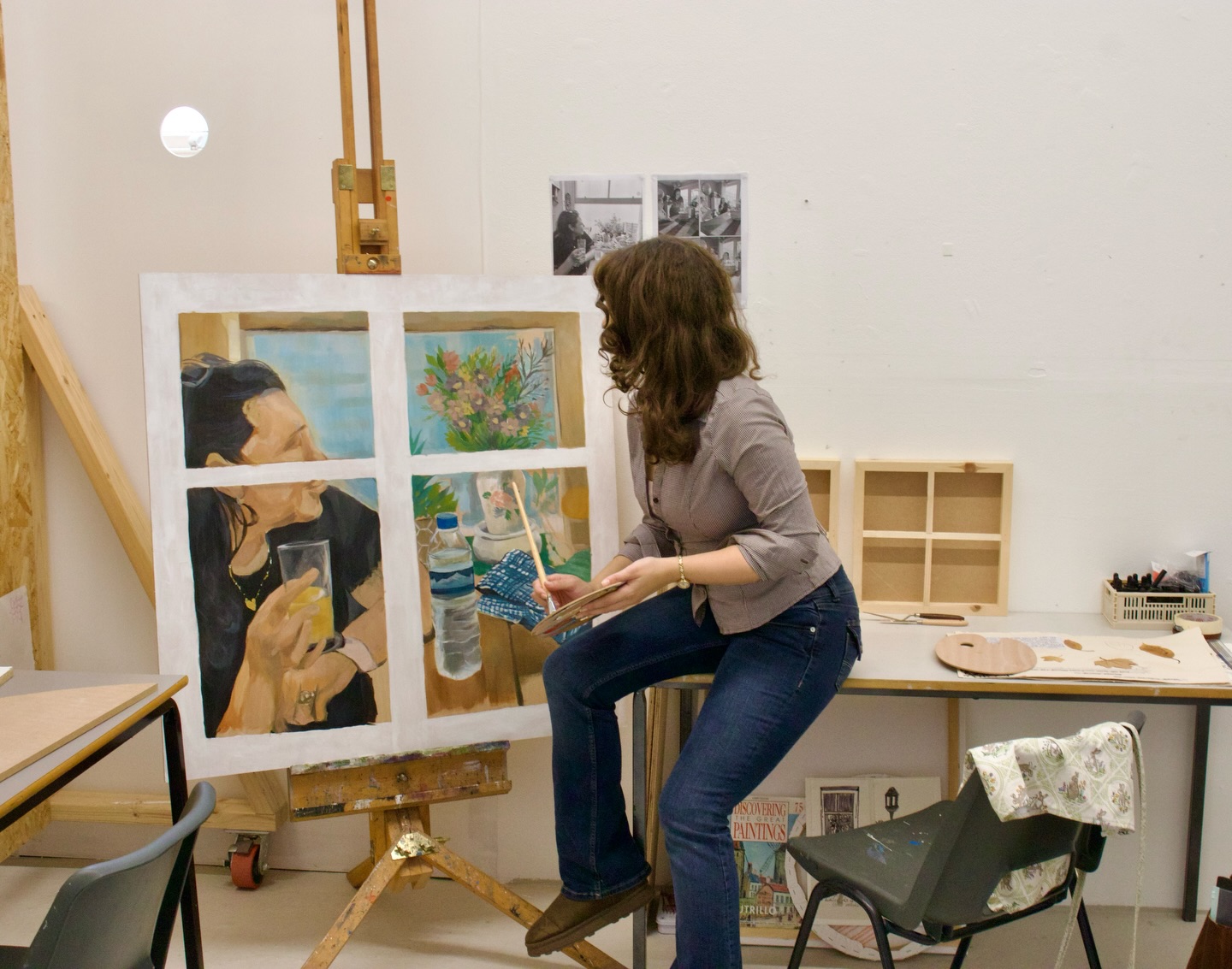 A woman sits on a stool in an art studio, painting a canvas on an easel. The canvas shows a person looking out a window. Art supplies and a chair are nearby, and black-and-white photos are pinned on the wall behind her.