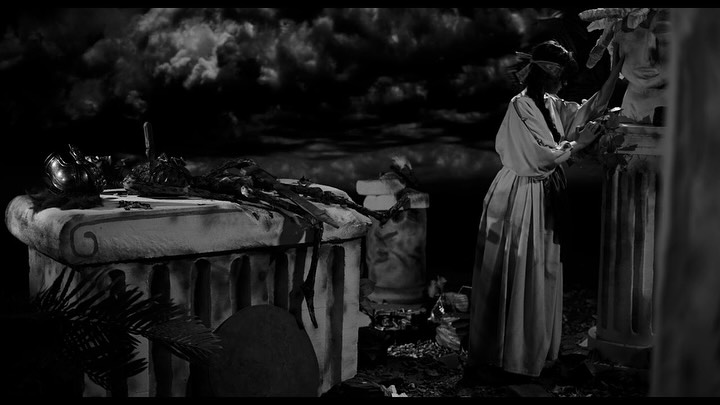 A woman in a long dress, blindfolded, touches a statue in a dark, dramatic setting with ancient ruins, scattered objects, and a cloudy night sky in the background.