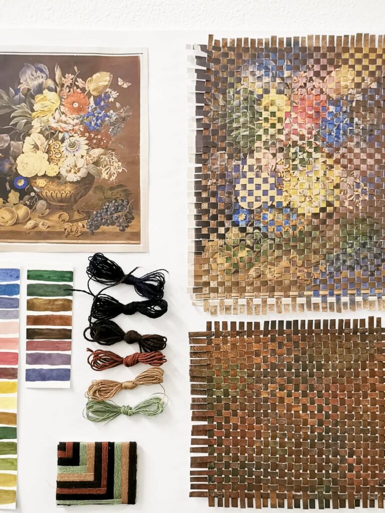 A wall display shows a floral still life print, a woven paper version of the artwork, color swatches, embroidery floss, and a small woven textile, all arranged neatly on a white background.