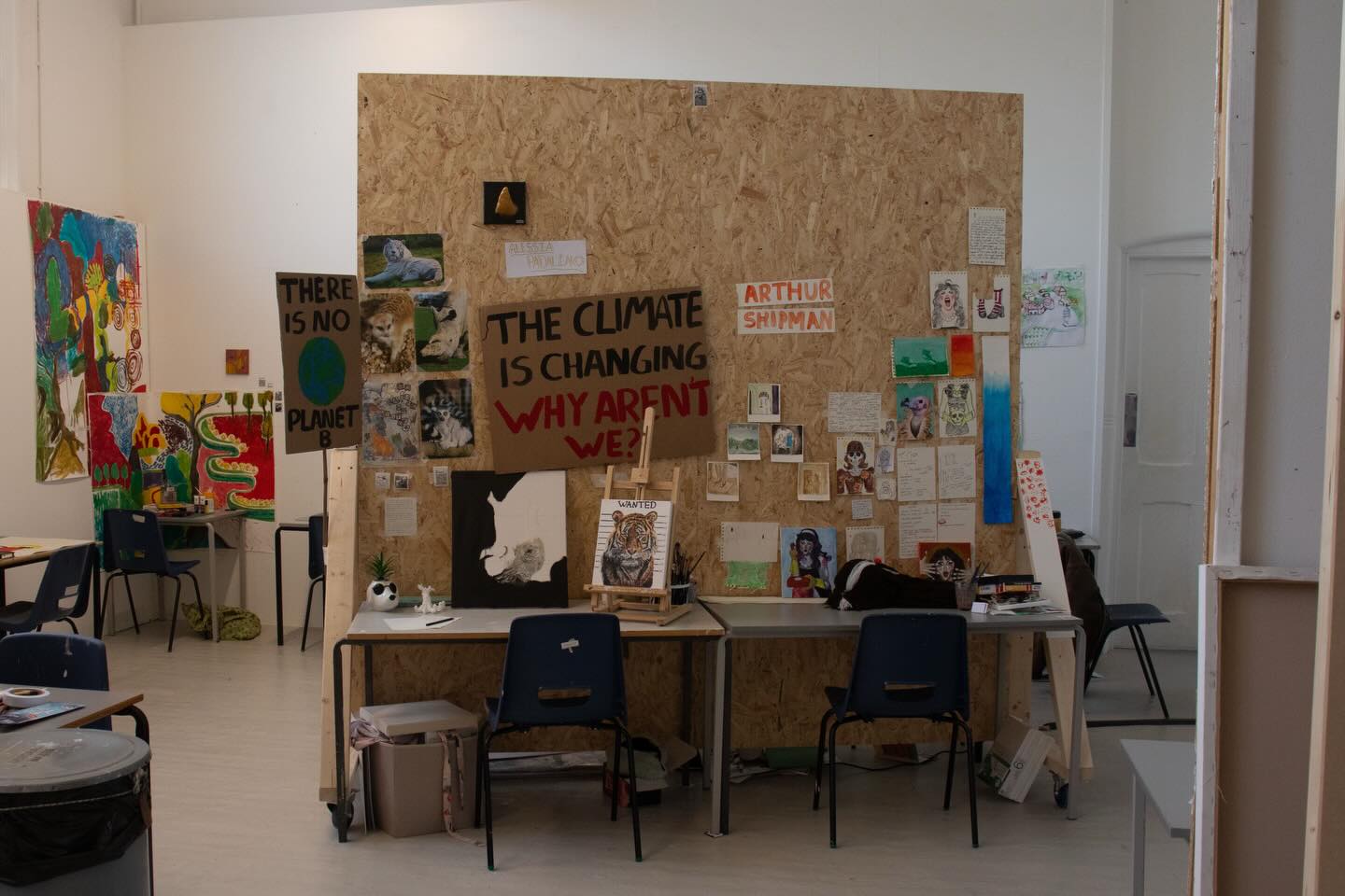 A classroom or art studio with posters and artwork on the walls, including a large sign reading "THE CLIMATE IS CHANGING WHY AREN'T WE?" and another saying "THERE IS NO PLANET B." Tables and chairs are scattered around the room.