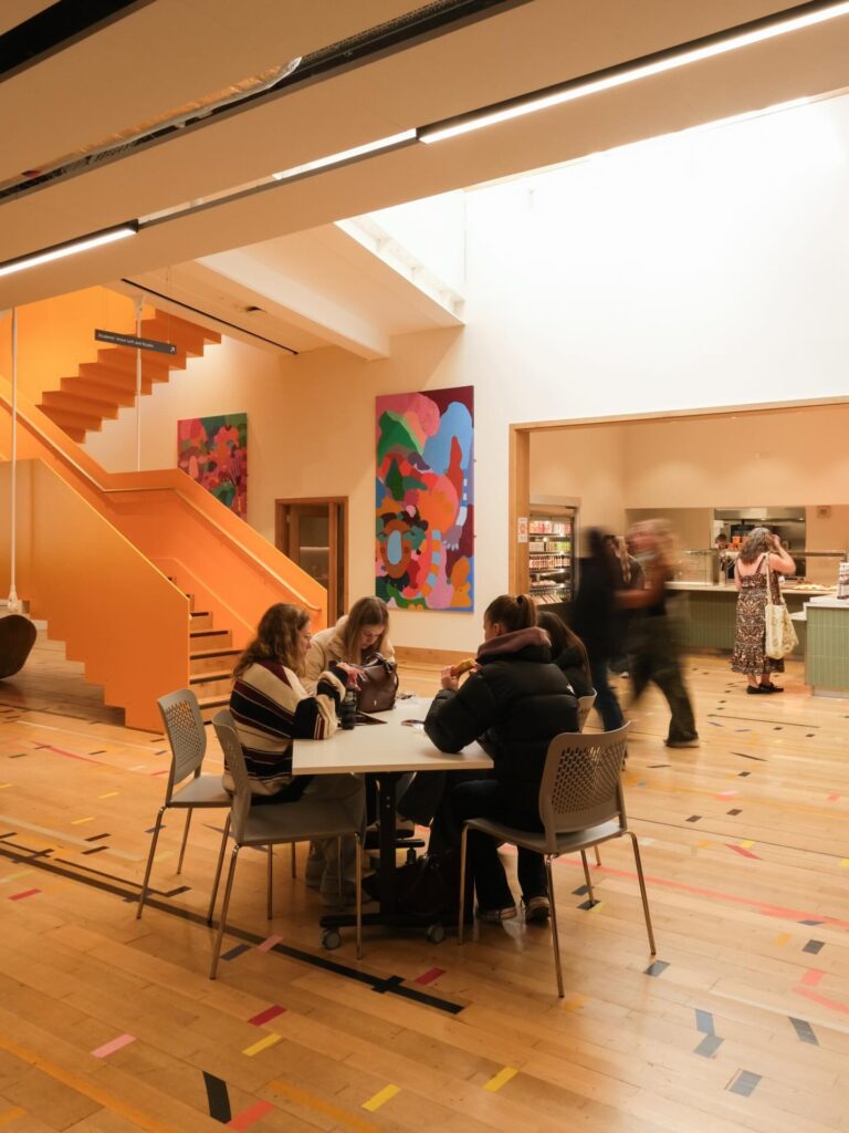 Three people sit at a round table in a brightly lit cafeteria with colorful abstract art on the walls, a wooden staircase, and a serving counter in the background. The floor has multicolored tape patterns.