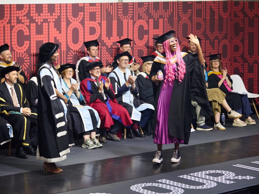 A graduate with long pink hair and a bright smile walks across the stage, waving, while faculty and audience members in academic robes clap and cheer during a graduation ceremony.