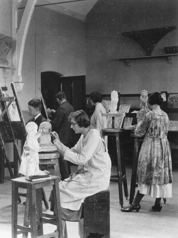 Several people work on sculptures and paintings in an art studio. One woman in the foreground sculpts a small statue, while others sculpt or paint at easels and tables in the background. The room has a high ceiling and art on display.