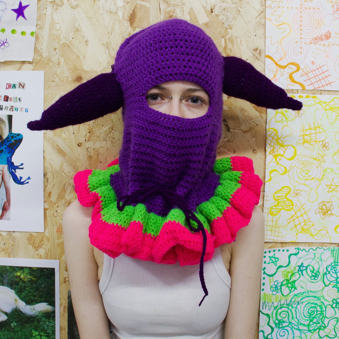 A person in a white tank top wears a purple crocheted mask with long pointed ears and a ruffled collar in neon pink and green, standing in front of a wall decorated with colorful drawings.