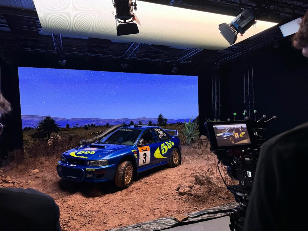 A blue rally car with yellow decals and the number 3 is parked on a rocky, dirt surface in front of a large screen displaying a nighttime landscape. Cameras and studio lights are visible in the setup.