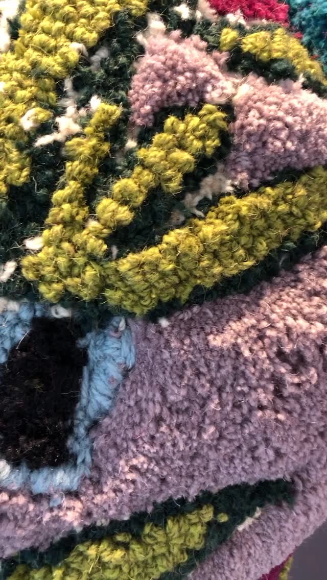 Close-up of a colorful, textured rug or tapestry with raised, fuzzy yarn in green, purple, yellow, blue, black, and cream, showing an abstract or floral pattern.