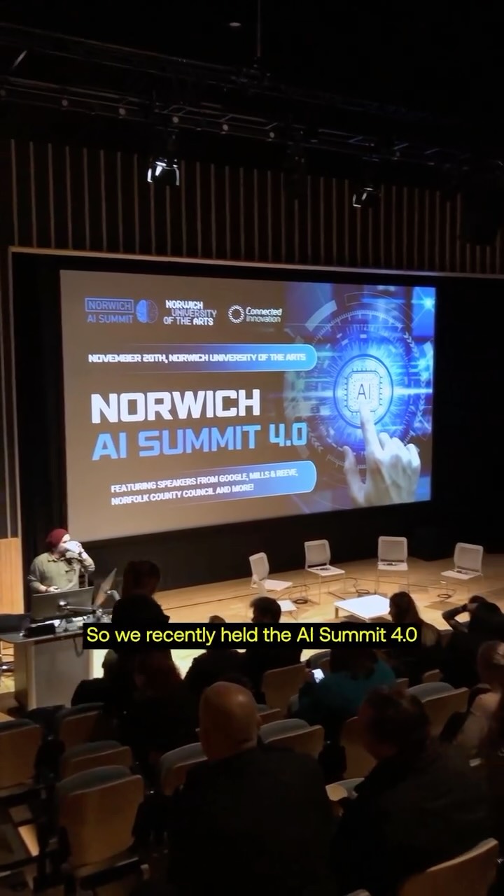 A large screen in an auditorium displays "Norwich AI Summit 4.0" with event details. Several people are seated, while one person stands at a podium. The subtitle reads, "So we recently held the AI Summit 4.0.
