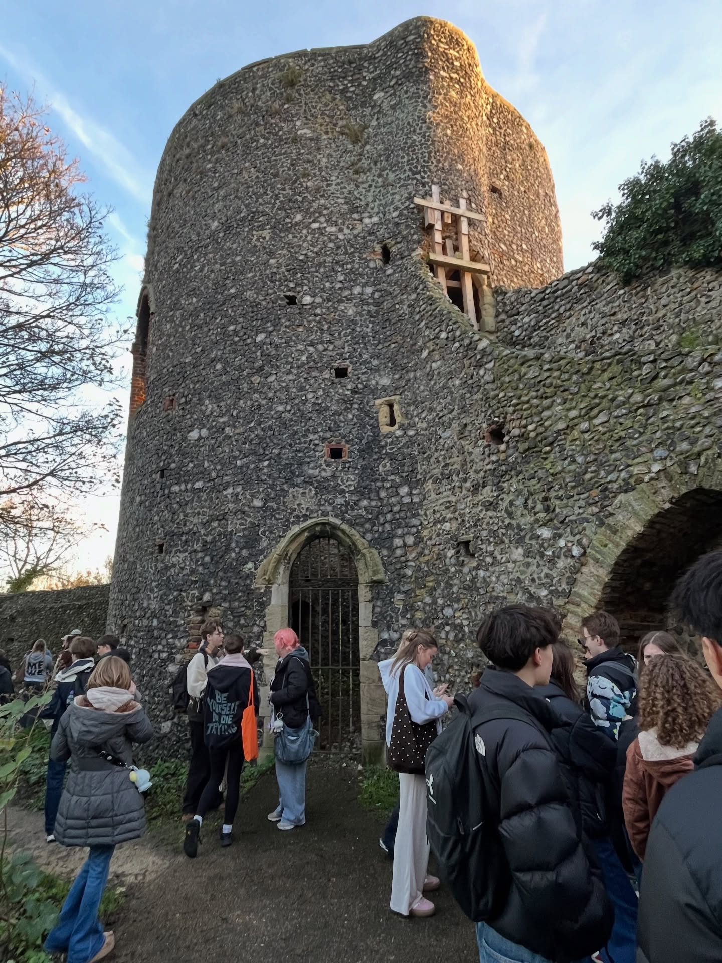 A group of people gather outside the entrance of an old stone tower with a rounded, weathered facade and small windows. The sky is clear and the scene appears to be during daylight.