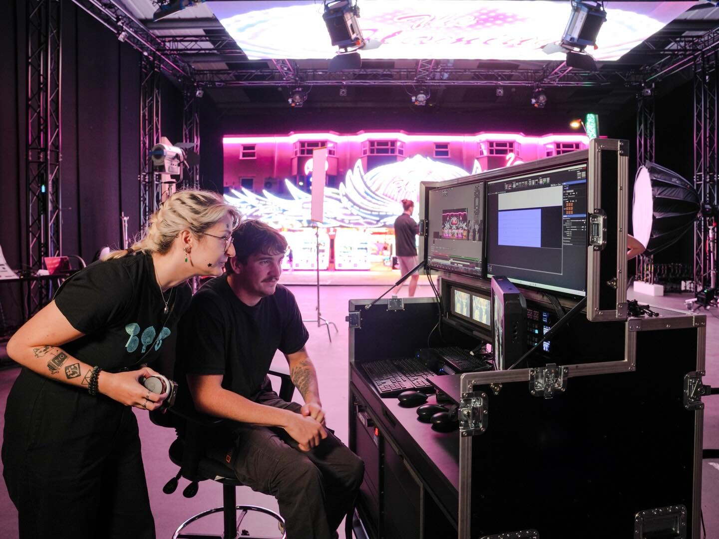 Two people collaborate in a studio, looking at computer monitors on a control desk. Bright stage lights and a large, colorful digital screen are visible in the background.