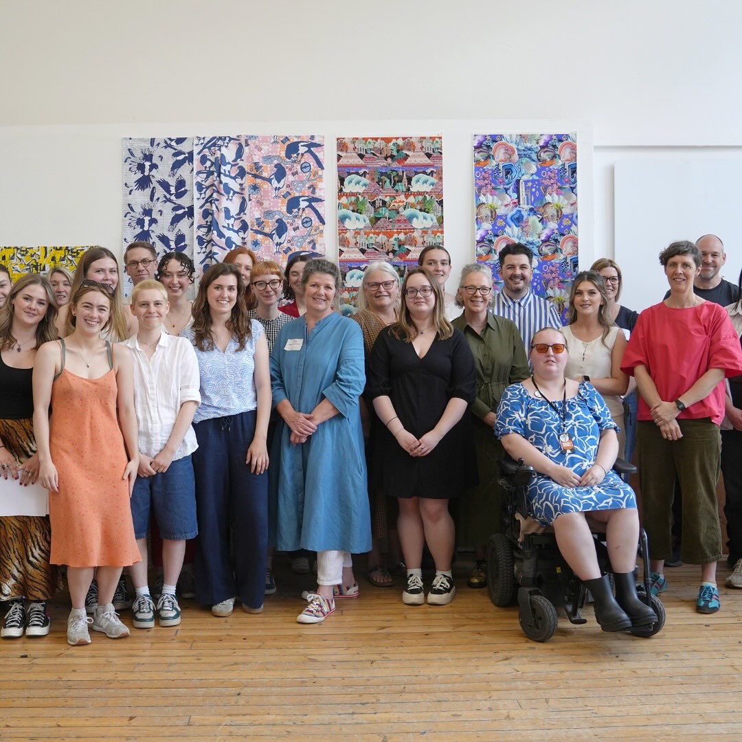 A group of people of various ages and genders pose together in front of colorful patterned artwork on a wall, standing on a wooden floor. One person is seated in a wheelchair at the front.